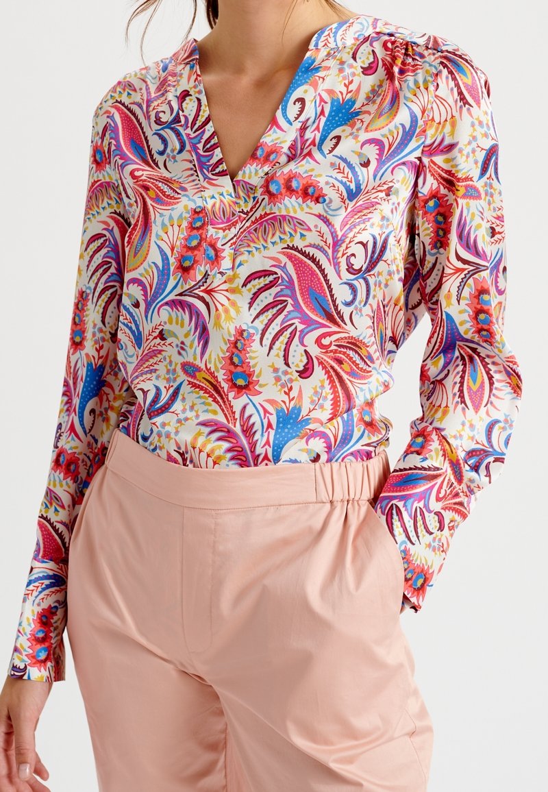 Femme portant un chemisier à motifs floraux colorés et cachemire avec un col en V, rentré dans un pantalon taille haute rose clair avec une ceinture élastique.