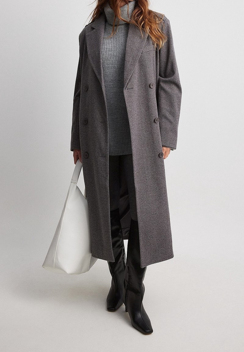 Femme portant un long manteau gris croisé sur un pull à col roulé gris, des bottes noires montantes jusqu'aux genoux, et tenant un grand sac à main blanc.
