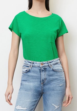 Nainen, joka pukeutunut kirkkaan vihreään lyhythihaiseen t-paitaan, jossa on rintatasku, ja korkeavyötäröisiin vaaleansinisiin kuluneisiin farkkuihin.