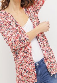 Stickad cardigan med ett flerfärgat mönster som främst är i röd, orange och vit. Korta ärmar, öppen front och en avslappnad passform.
