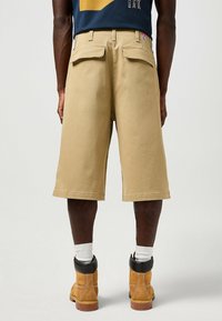 Bermudas cargo beige con un corte suelto, dos bolsillos traseros y detalles de costura visibles. Combinadas con botas marrones y calcetines blancos de caña corta.