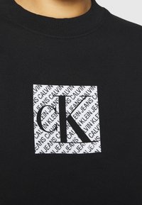 Sudadera negra con un logotipo rectangular blanco que presenta el texto repetido "CALVIN KLEIN JEANS" y un destacado "CK" en negrita negra.