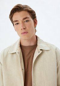 Beige denimmik Jacket, jolla on kaulus, napit ja tekstuoitu pinta, päälle puettuna ruskea ribbipaita. Vaalea tausta.