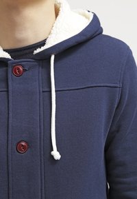 Sudadera con capucha azul marino con una capucha forrada de forro polar blanco, que cuenta con botones rojos, cordones ajustables y una tela texturizada para proporcionar calidez y comodidad.