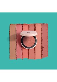 Poudre blush compacte avec un couvercle rose et un motif en relief. Des échantillons autour montrent des nuances de mauve et de pêche sur un fond teal.