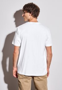 Bílý bavlněný tričko s krátkým rukávem a kulatým výstřihem, hladká textura a uvolněný střih, doplněné béžovými kalhotami.