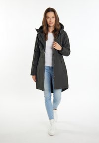Lang sort vandtæt jakke med hætte, lynlåslukning foran, justerbar talje og to sidelommer, båret over en hvid skjorte og blå jeans.