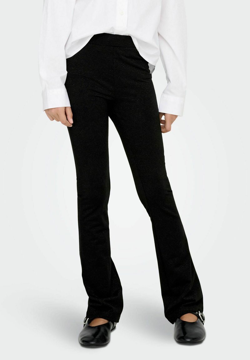 Osoba ubrana w czarne dopasowane spodnie w fasonie dzwonów, czarne płaskie buty oraz białą koszulę z długim rękawem zapinaną na guziki, na jednolitym tle.