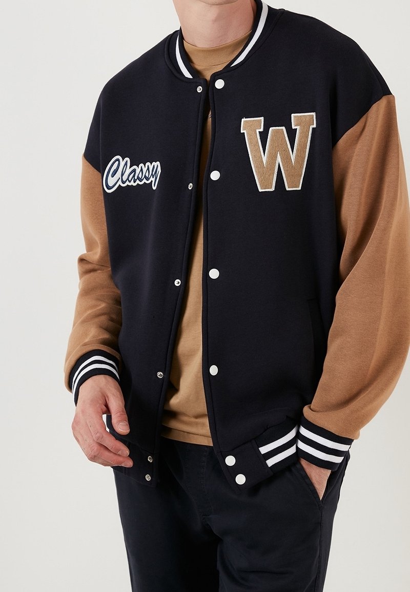 Svart och brun varsityjacka med randiga muddar, broderad med "Classy" och en stor "W"-logotyp. Framknäppning och sidofickor.