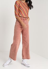 Femme portant un jean large rose, un haut à motifs losanges multicolores et des baskets montantes blanches, debout devant un fond uni.
