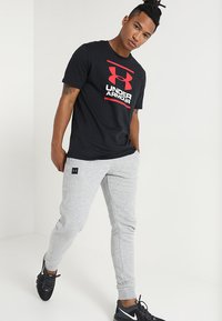 Camiseta de algodón negra con el logo rojo de "Under Armour", combinada con pantalones de chándal grises, que presentan un diseño ajustado. Zapatillas deportivas negras completan el look.