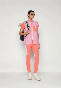 Rozā un koralja sportiskais tērps ar bezpiedurkņu bezgalīgu vestu un pieguļošām legingām, apvienots ar mugursomu un sporta apaviem. Strukturēti materiāli un krāsu blokēšana.