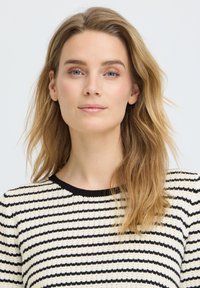 Femme blonde aux yeux bleus portant un pull rayé noir et blanc, face à l'avant avec une expression neutre et les cheveux lâchés.