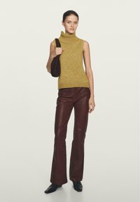 Maglione a collo alto in senape, abbinato a pantaloni svasati in ecopelle bordeaux. La modella tiene in mano una borsa a spalla in tessuto marrone.