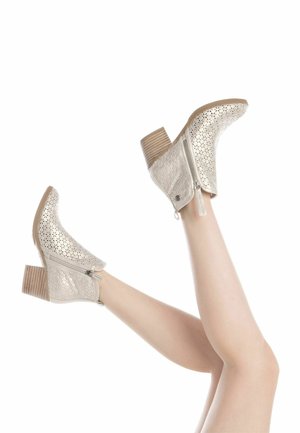 Botas de tobillo plateadas metálicas con patrón perforado, cremalleras laterales y tacones de madera apilados. La forma es ligeramente curva en la parte superior.