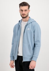 Felpa con zip di colore azzurro chiaro, con cappuccio regolabile tramite cordino, tasche frontali e un piccolo logo bianco sul petto. Tessuto morbido e strutturato.