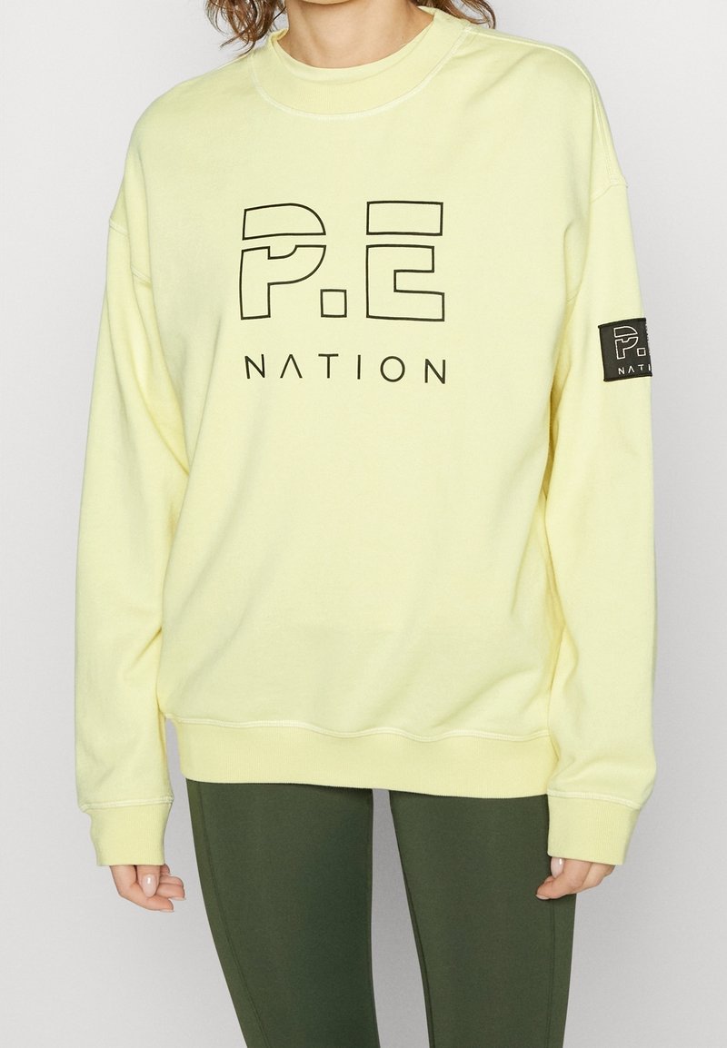 Sweat-shirt jaune clair avec logo noir "P.E NATION", col rond côtelé et manches avec poignets. Dispose d'un patch de marque sur la manche gauche.