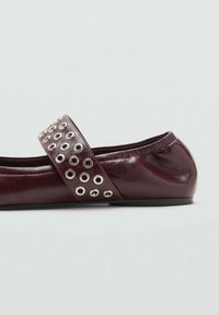 Burgundy nahkainen tasapohjainen kenkä, jossa on pyöreä varvas. Kenkässä on leveä hihna, joka on koristeltu hopeanvärisillä metallilenkeillä yksityiskohtien lisäämiseksi.