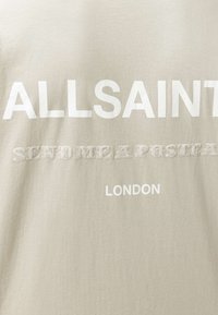 Μπεζ ύφασμα με λευκό κείμενο: «ALLSAINT», υφασμένα γράμματα που λένε «ΣΤΕΙΛΕ ΜΟΥ ΜΙΑ ΚΑΡΤΟΛΙΝΑ» και «LONDON» τυπωμένο από κάτω.