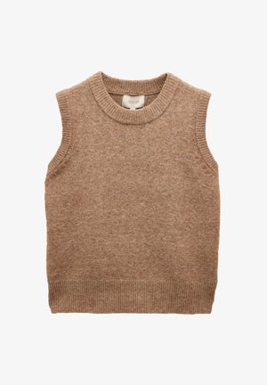 Junge Frau mit glatten braunen Haaren, die ein ärmelloses braunes Stricktop und weiße Hose mit Kordelzug trägt, steht vor einem einfarbigen beigen Hintergrund.