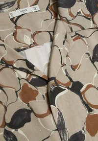 Chemise à manches longues en tissu léger, présentant un fond beige avec des motifs abstraits marron, noir et crème ainsi qu'un col de chemise.