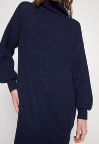 Stickad klänning i mörk marinblått med en texturerad yta, med hög hals och vida ärmar, vilket ger en lös och avslappnad passform.