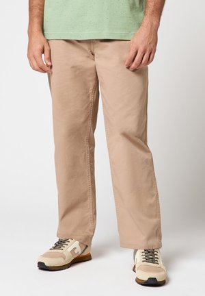 Homme portant un pantalon beige droit, un t-shirt vert tacheté, et des baskets beiges avec des accents blancs et marron, debout sur un fond blanc.