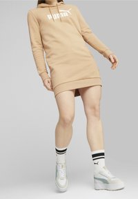 Krémové mikinové šaty s velkým bílým logem "PUMA". Obsahují šňůrkovou kapuci a žebrované manžety. V páru s bílými teniskami a pruhovanými ponožkami.