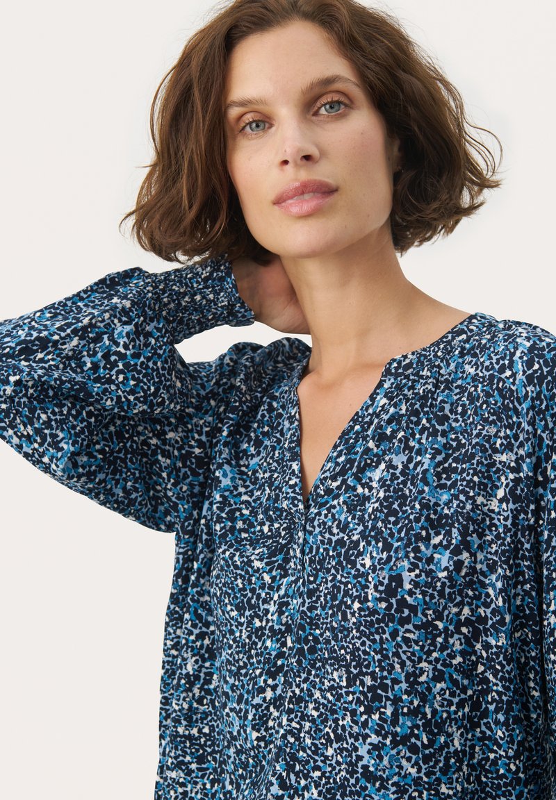 Bluse mit blauem Muster, V-Ausschnitt und gerafften Ärmeln. Hergestellt aus leichtem Stoff, der eine Mischung aus dunklen und hellen Blautönen zeigt.