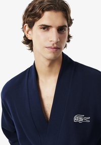 Accappatoio blu navy realizzato in tessuto testurizzato, con colletto a scialle e ricamo del logo Lacoste sul lato sinistro del petto.