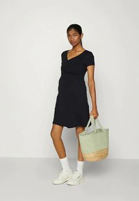 Mulher vestindo um vestido preto em V e ténis brancos, segurando uma bolsa de palha bicolor, de pé contra um fundo claro e liso.