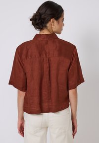 Rust-colored linen short-sleeve shirt with a back pleat, straight hem, and pointed collar, worn with white denim pants.