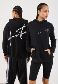 Zwei Personen mit geflochtenem Haar tragen schwarze Hoodies mit dem Schriftzug "Deine Zukunft"; einer blickt nach vorne mit den Händen in den Taschen, der andere blickt nach hinten.