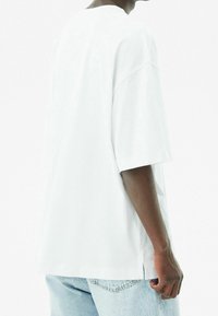 T-shirt bianco oversize con maniche corte, scollo rotondo, spacchi laterali e texture in cotone morbido. Indossato con jeans blu chiaro.