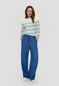 Gestreiftes Oberteil mit Bootsausschnitt und 3/4-Ärmeln; blaue Denim-Hose mit weitem Bein und Kordelzugbund; getragen mit weißen Sneakers. Neutraler Hintergrund.