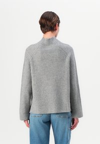 Grå strikkegenser med høy hals og ribbestrikket tekstur, med lange, vide ermer og en avslappet passform. Brukt sammen med lyseblå jeans.