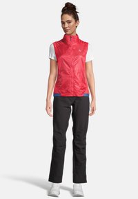 Rote ärmellose Jacke mit hohem Kragen, ausgestattet mit einem Reißverschluss auf der Vorderseite und kontrastierenden blauen Akzenten am Saum, kombiniert mit schwarzen Hosen und weißen Schuhen.