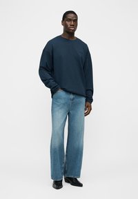 Bărbat purtând o cămașă cu mânecă lungă bleumarin, blugi largi, de culoare albastru deschis, și cizme negre cu vârf ascuțit, stând pe un fundal simplu.