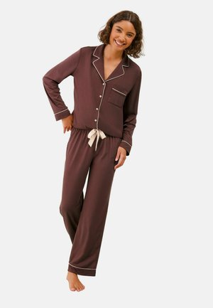 Smiling woman with curly hair wearing brown long-sleeve buttoned pajama top and matching pants with white trim and tie waist, standing barefoot.