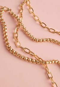 Zelta ķēde, kas sastāv no savienotiem ovāliem un apaļiem segmentiem ar dažādām tekstūrām un spīdīgu apdari, izlikta uz maigi rozā fona.