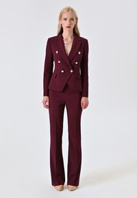 Blazer doppiopetto bordeaux con bottoni dorati, abbinato a pantaloni coordinati. Tessuto testurizzato; design aderente, con scollatura a V profonda.