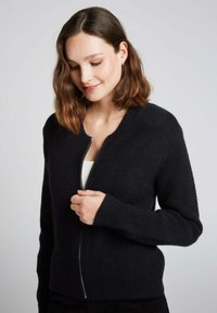 Cardigan zippé noir avec une texture côtelée, un col rond et des manches longues. Confectionné dans un matériau doux, il présente des détails de design minimalistes.