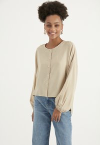 Beige Bluse mit Knöpfen, runder Halslinie, langen Puffärmel und cropped Saum. Getragen mit hellblauen Jeans. Glatte Stofftextur.