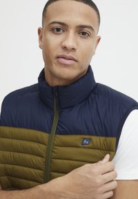 Polstret vest med en navy top og olivengrøn bund, høj krave og frontlynlås. Har et lille logo på brystet.