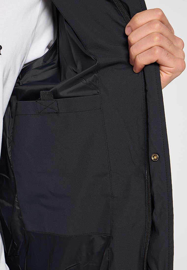 Interior de una chaqueta negra que muestra una tela lisa y forrada con un bolsillo oculto y un pequeño lazo para sujeción o enganche. Material duradero.