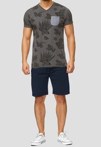 T-shirt gris à col en V avec imprimé floral noir et poche bleue, associé à un short marine et des baskets blanches. Coupe décontractée et en coton.