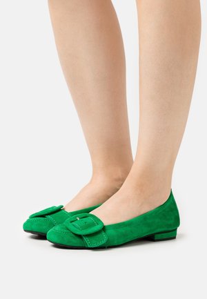 Voeten in felgroene suède ballerina's met grote vierkante gesp, staand op een witte ondergrond.