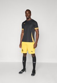 Svart treningstrøye med gullakenter, gule shorts med logo, svarte sokker og svarte fotballsko, som viser et sporty antrekk.