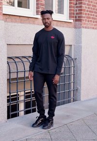 Black long-sleeve shirt with red logo, paired with black sweatpants and black sneakers. Features sleek design and smooth texture.