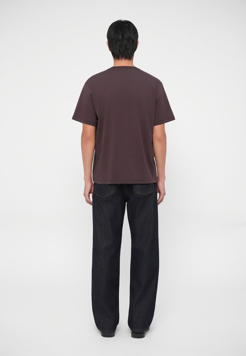 Donkerpaarse katoenen T-shirt met korte mouwen, gedragen met losse donkerdenim jeans, gezien van achter tegen een witte achtergrond.